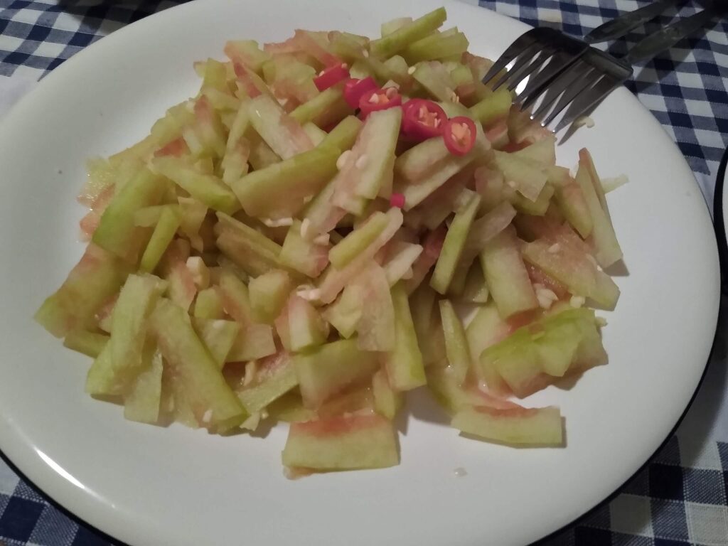 A white plate holds a pile of thinly sliced watermelon rind pieces, with a few slices of red chili pepper on top. The plate is on a blue and white checkered tablecloth, with a fork and knife placed nearby.