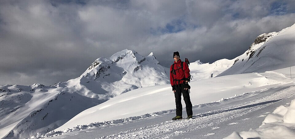 A person dressed in a red jacket and winter hiking gear stands on a snowy trail, surrounded by pristine, snow-covered mountains under a cloudy sky. The terrain is smooth with deep snowbanks, and the peaks loom dramatically in the background.