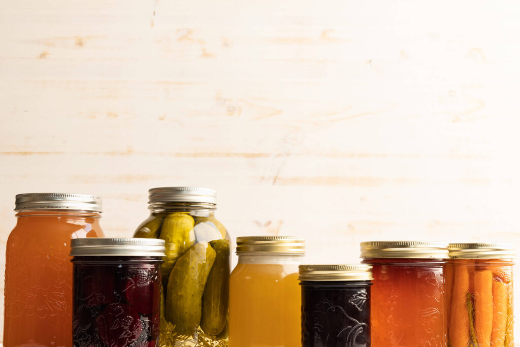 Border of glass jars, pickled beets, pickles, filled carrots, honey and apple jelly, in autumn colours on a white wood background with copy space