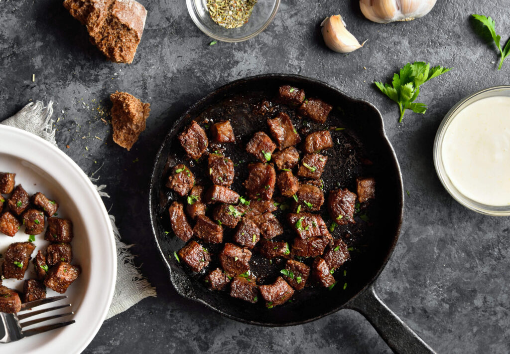 Garlic butter steak bites in frying pan over dark stone background. Top view, flat lay