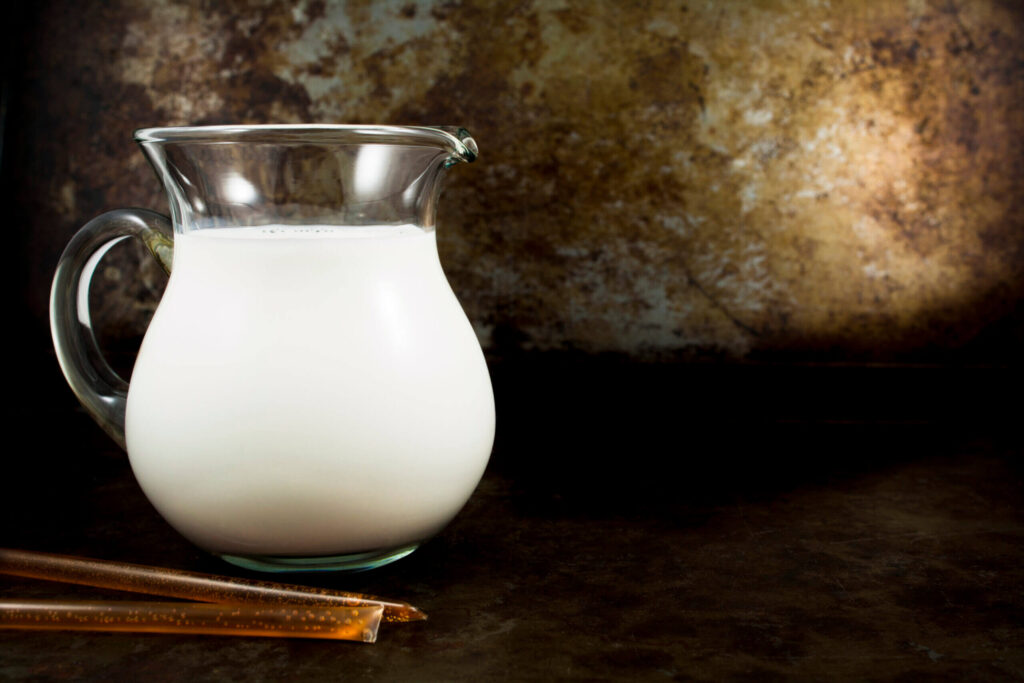 Glass pitcher of milk and honey sticks against a rustic backdrop