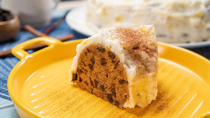 A slice of spiced fruitcake with raisins and dark fruit pieces, covered in a creamy white frosting and dusted with cinnamon. The cake sits on a vibrant yellow plate, with the rest of the cake visible in the blurred background. Cinnamon sticks and a warm-toned setting suggest a cozy, autumnal or holiday vibe.