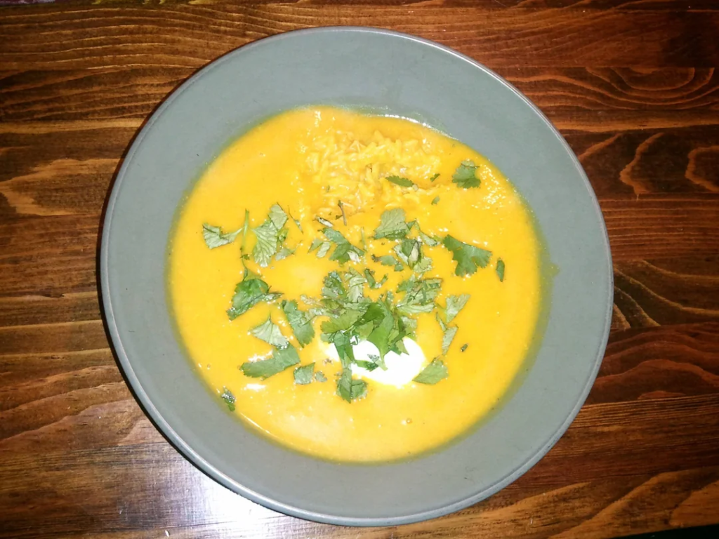 A bowl of creamy orange soup, possibly a spiced carrot or squash soup, garnished with fresh chopped cilantro and a dollop of sour cream or yogurt. The dish also contains a portion of rice, partially submerged in the soup. Served in a muted gray bowl on a rustic wooden table.