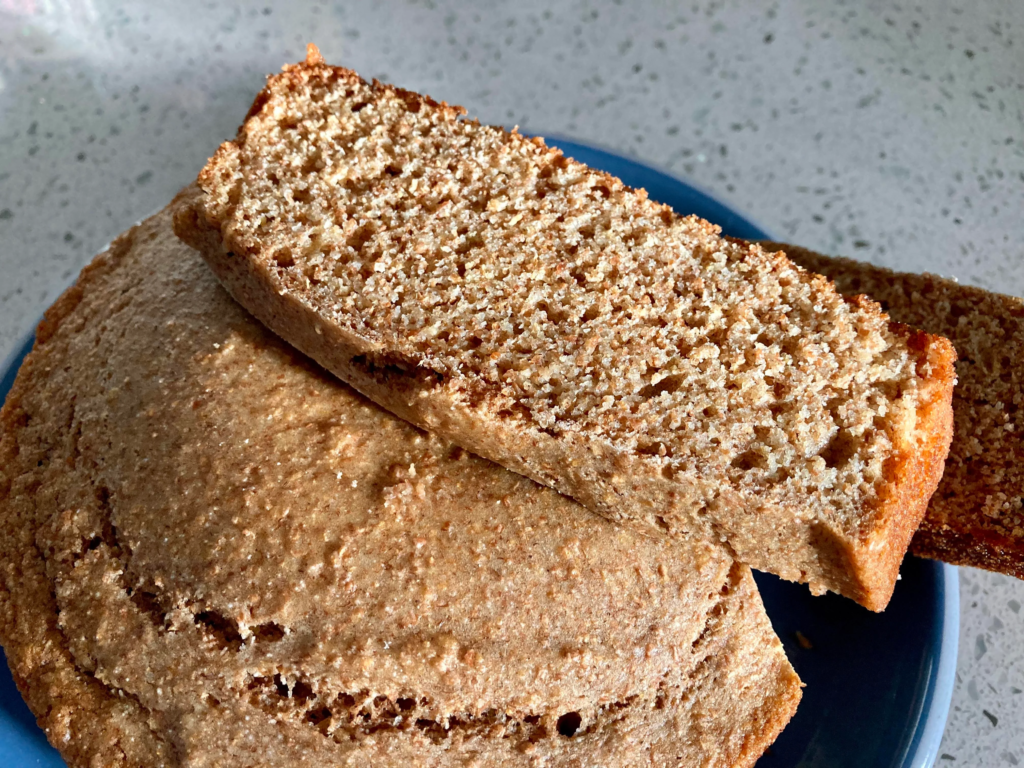 A close-up of freshly baked whole wheat or spiced bread, with a rustic golden-brown crust and a soft, airy interior. One slice is resting on top of the loaf, showing its crumb texture and slightly rough surface. The bread sits on a simple blue plate, with natural light highlighting its homemade appearance.