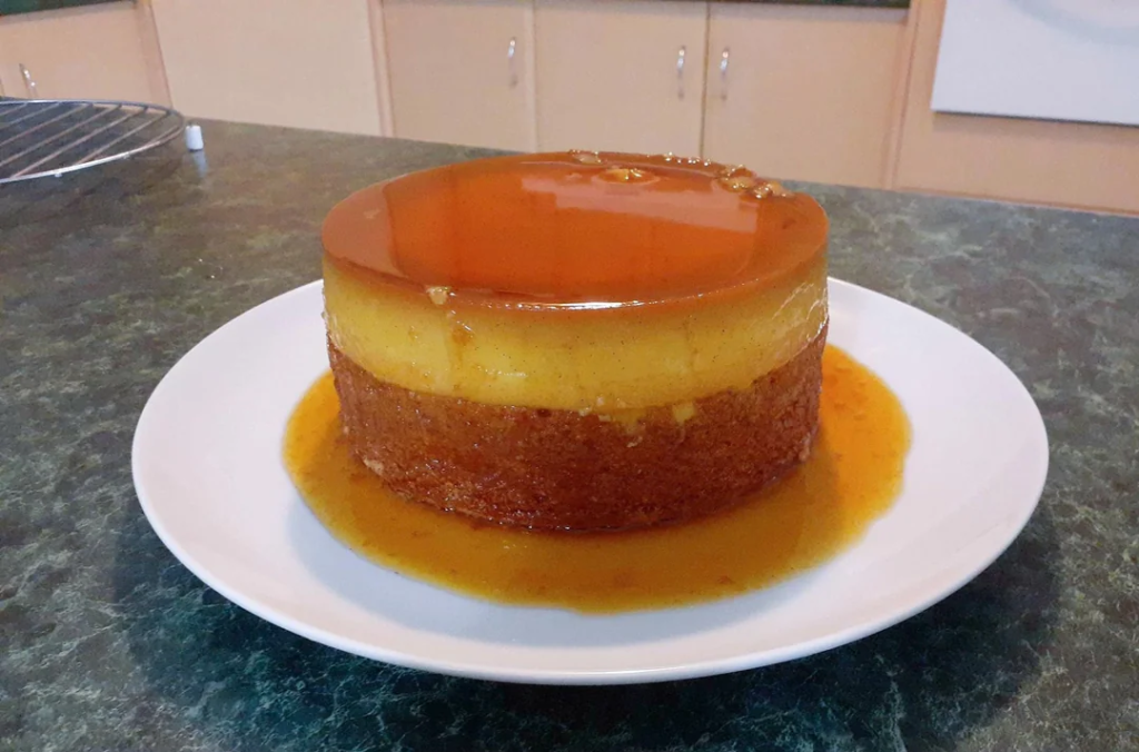 A beautifully layered chocoflan, also known as magic cake, served on a white plate. The dessert features a rich caramel glaze on top, a smooth flan layer in the middle, and a moist chocolate cake base, with caramel sauce pooling around the plate. The background shows a home kitchen with cabinets and a countertop.