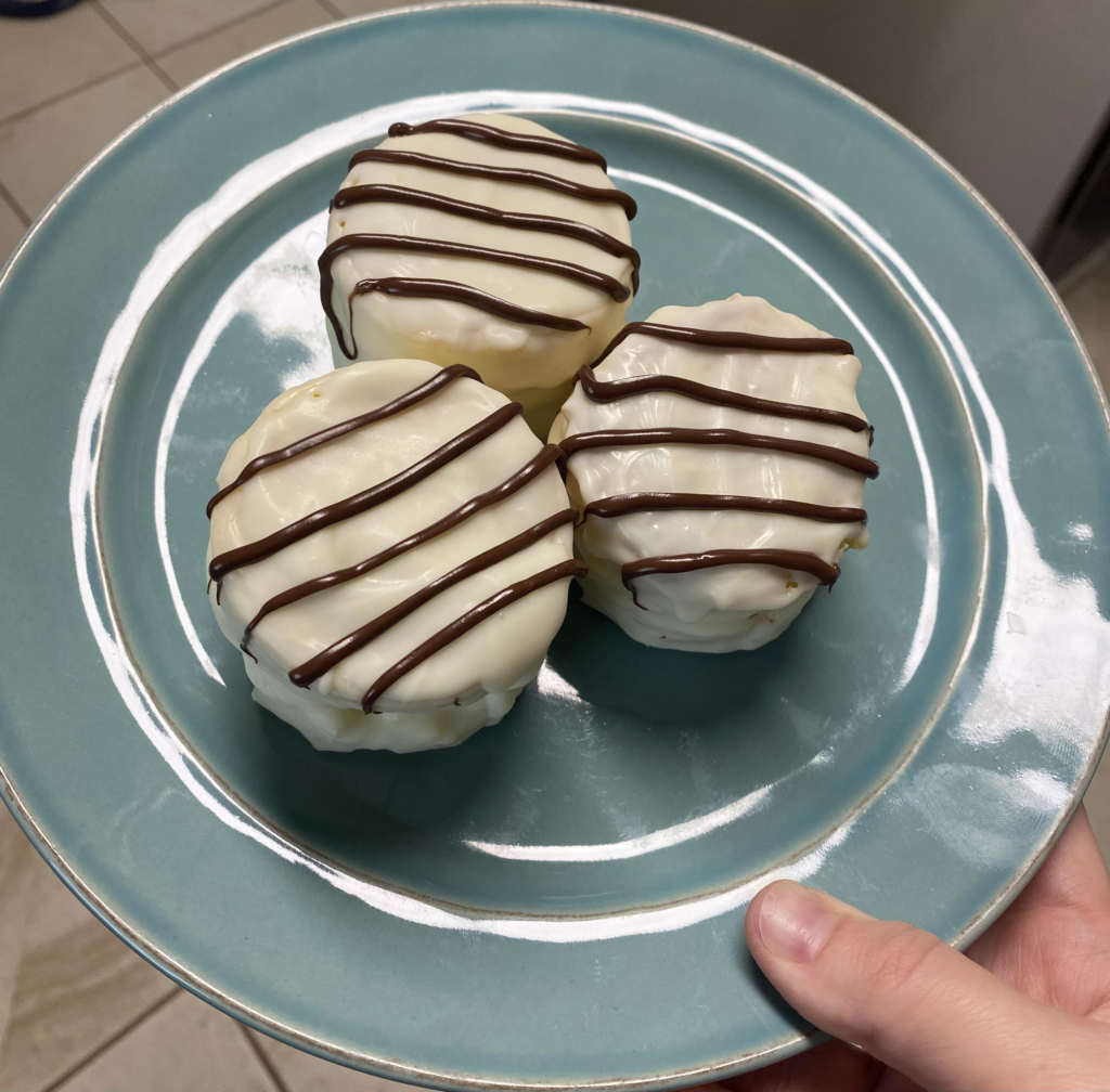 Three small, round desserts coated in smooth white chocolate and drizzled with dark chocolate stripes, arranged on a blue plate. The treats have a uniform shape with a slightly glossy finish, suggesting a creamy or cake-like filling inside. A hand is holding the plate, with a kitchen setting visible in the background.