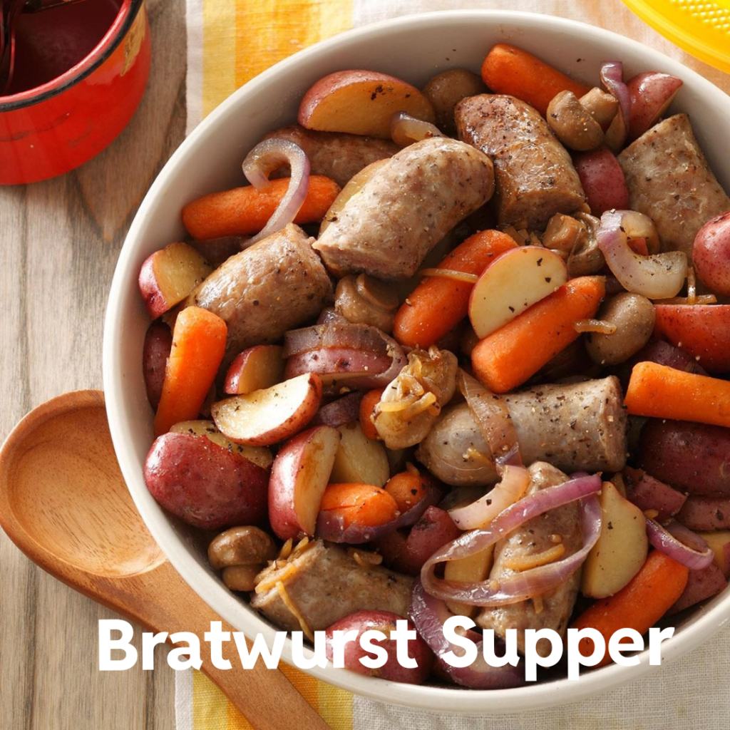 A hearty bowl filled with roasted bratwurst sausages, sliced red potatoes, baby carrots, mushrooms, and thinly sliced red onions. The dish is seasoned with black pepper and herbs, with a rustic wooden spoon beside it. Bold white text reading "Bratwurst Supper" overlays the bottom of the image.