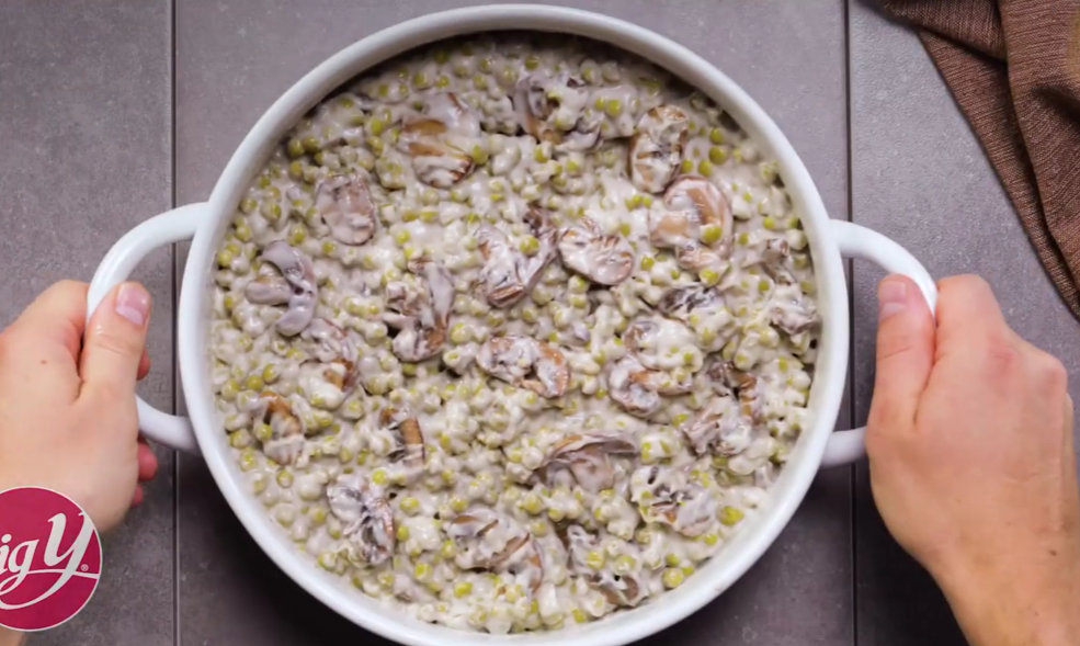 A large white casserole dish filled with a creamy mushroom and pea mixture, possibly a classic side dish or casserole. The dish is thick and hearty, with visible chunks of sliced mushrooms and green peas in a white sauce. Two hands are holding the dish on a tiled countertop, with a brown cloth napkin in the background. The bottom left corner features a "Big Y" logo.