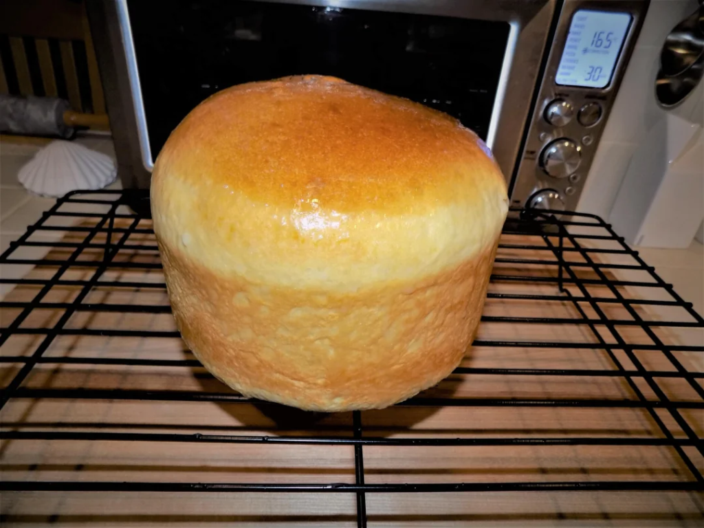 A freshly baked, tall loaf of bread with a golden-brown, glossy crust cooling on a black wire rack. The bread has a smooth, domed top and appears to have been baked in a cylindrical bread machine. In the background, a toaster oven with a digital display and kitchen utensils are visible on the countertop.