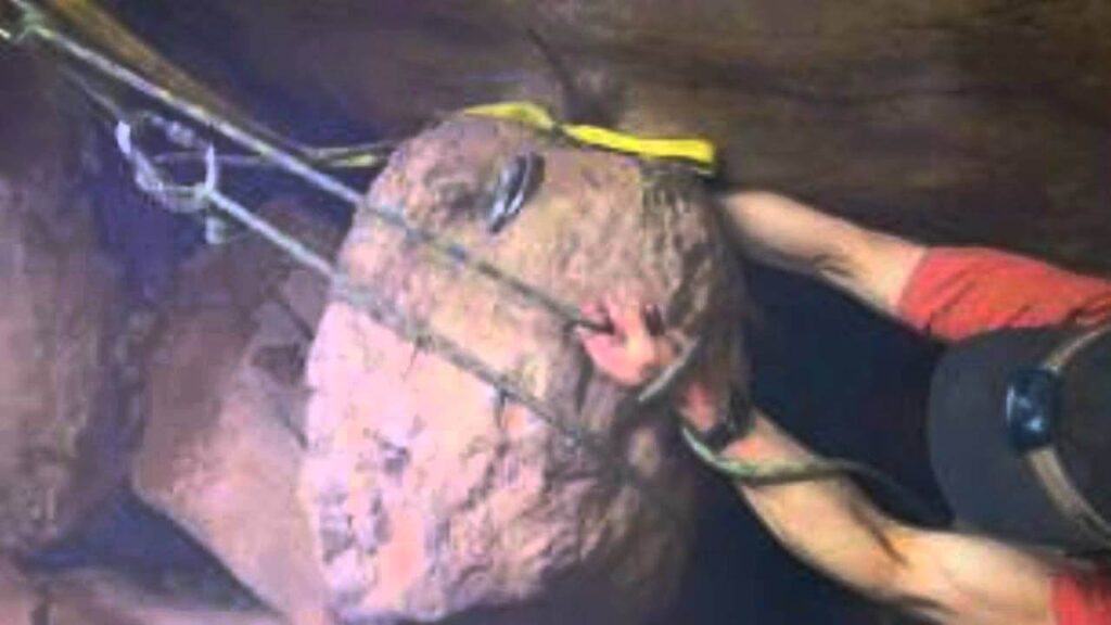 A man wearing a headlamp and red shirt is trapped in a narrow canyon by a large boulder, with climbing ropes and gear visible securing the rock. His right arm is pinned, and the scene appears tense and confined within a dimly lit crevice.