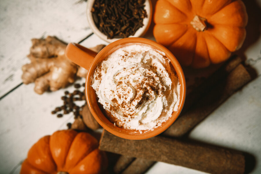 An Autumn favorite hot drink, the pumpkin spice flavored coffee, tea, or chai, complete with fresh pumpkins and whipped cream topping. Cinnamon sticks, ginger, cloves, and allspice surround the mug of spicy warm goodness. Rustic wood table.